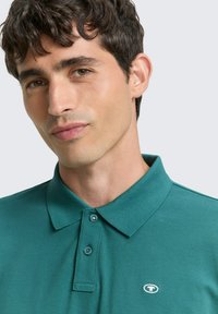 Türkisfarbenes Poloshirt aus strukturiertem Stoff, mit Kragen, zwei Knöpfen und einem kleinen Logo auf der linken Brustseite.