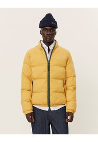 Geel pufferjack met een hoge kraag, ritssluiting aan de voorkant en grijze accenten; gecombineerd met een marineblauwe gebreide beanie en een wit shirt eronder.