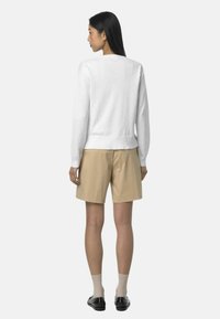 Maglione bianco a collo alto con maniche lunghe, polsini e orlo a coste, abbinato a pantaloni corti color khaki. I calzini sono chiari e le scarpe sono nere.