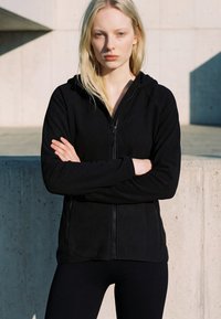 Mlada žena plave kose u crnoj majici s kapuljačom na patent, stoji vani s prekriženim rukama ispred betonskog zida.