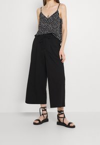 Camisola a pois in bianco e nero con spalline sottili abbinata a pantaloni ampi neri e sandali neri con fasce.
