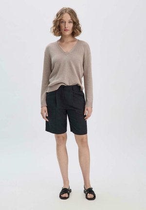 Beige gebreide trui met een V-hals en lange mouwen, gecombineerd met zwarte shorts tot de knie. Model draagt zwarte sandalen.