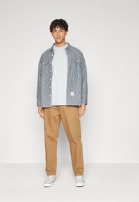 Stribet blå og hvid skjorte med knapper over en grå t-shirt, parret med lysebrune bukser og grå sneakers på en ensfarvet baggrund.