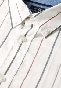 Valkoinen napillinen paita, jossa on monivärisiä pystysuoria raitoja punaisena, sinisenä ja mustana. Paidassa on klassinen kauluskotelointi ja valkoiset napit.