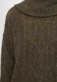 Pull en maille vert foncé avec de gros motifs torsadés et un grand col roulé plié.