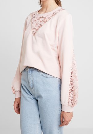 Vaaleanpunainen collegepaita, jossa on pitsikoristelua kaula-aukossa kolmionmuotoisena ja pitsiyksityiskohtia hihoissa. Kevyt kangas, rento istuvuus.