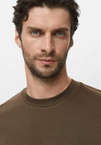 Smeđi pamučni majica kratkih rukava s ravnim izrezom, rebrastim okovratnikom, glatke teksture i uskog kroja, nosi muškarac svijetloplavih očiju i bradom.