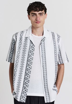 Un jeune homme aux cheveux courts et bouclés de couleur sombre, portant une chemise blanche avec des rayures géométriques verticales noires, les mains dans les poches, devant un fond clair uni.