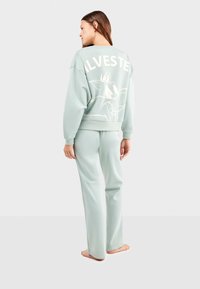 Lichtgroene sweatshirt en bijpassende broek hebben een grote grafische afbeelding van een cartoonfiguur op de achterkant. Zacht, casual stof met een relaxte pasvorm.