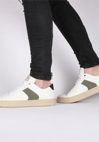 Zapatillas de cuero blancas con detalles verdes, parte superior texturizada y suela de goma beige. Presentan un diseño con cordones y una pestaña trasera negra.