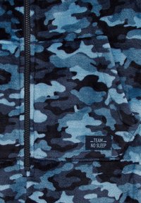 Polārā jaka ar zilu un melnu kamuflāžas rakstu, ar rāvējslēdzēju, kā arī priekšējo kangaroo kabatu. Etiķete norāda "TEAM NO SLEEP". Mīksta tekstūra.