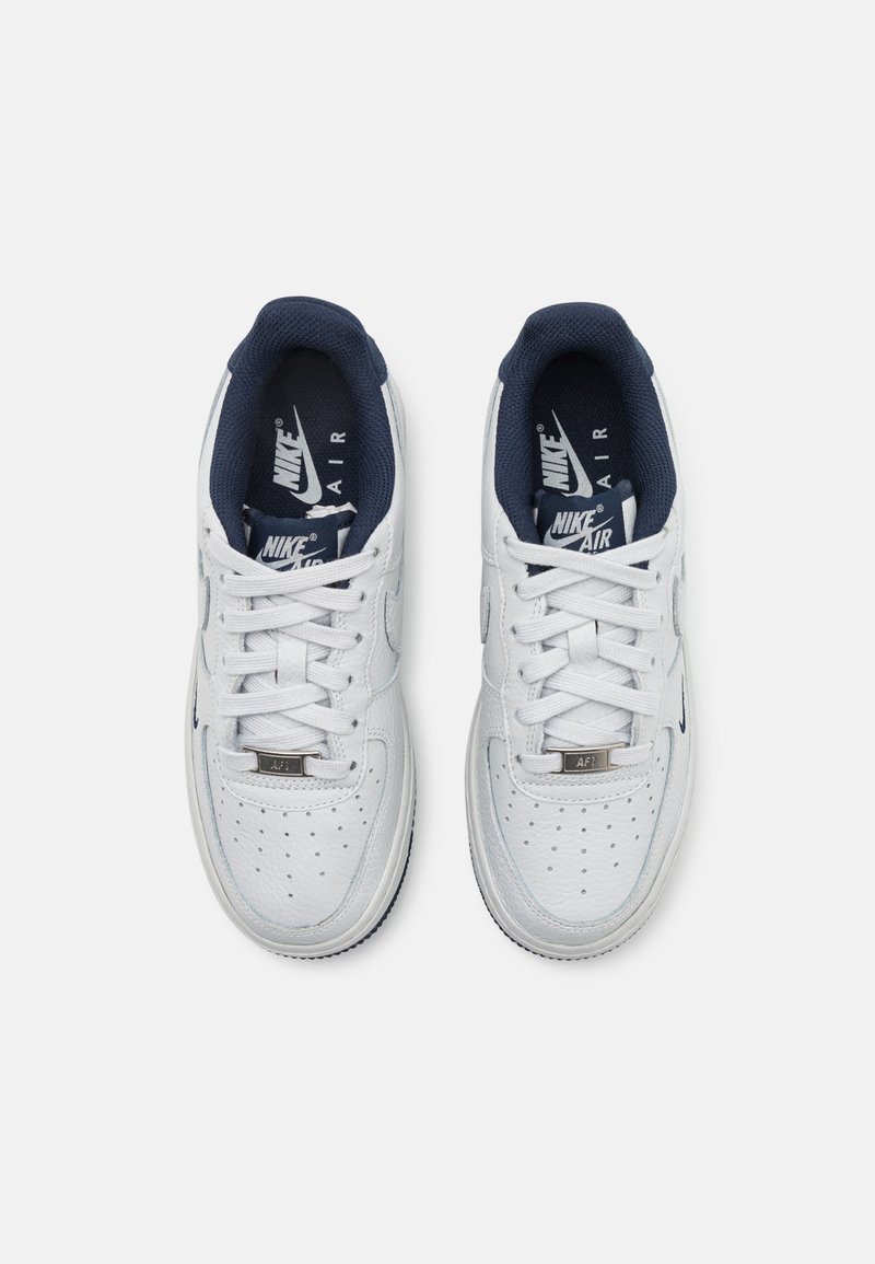 Zapatillas blancas de cuero con detalles azules, que presentan perforaciones, cordones planos y un logo en la lengüeta. La suela es blanca con un patrón texturizado.