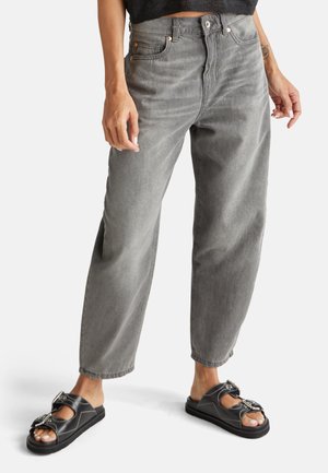Person trägt lockere graue Jeans und schwarze Sandalen mit Schnalle steht vor einem schlichten hellen Hintergrund.
