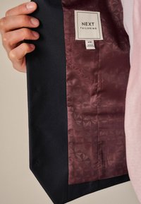 Intérieur d'une veste tailleur marine avec une doublure en tissu texturé noir et un panneau en satin bordeaux présentant un motif géométrique. Étiquette de marque visible.