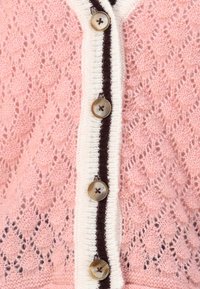 Adīta džemperis ar rozā mežģīņu rakstu, ar krēmkrāsas joslu un tumši brūniem akcentiem, kā arī lielām pogām ar krusta dizainu.