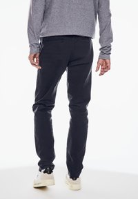 Tmavě modré bavlněné kalhoty s úzkým střihem a zadními kapsami, doplněné bílými a černými teniskami, viděné zezadu.