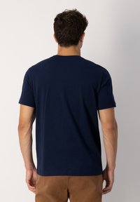 Muž s tmavými kudrnatými vlasy, na sobě má jednoduché tmavě modré tričko s krátkým rukávem a hnědé kalhoty, otočený zády k jednoduchému světlému pozadí.