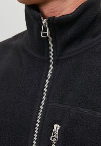 Jachetă din fleece neagră cu guler înalt, dotată cu un fermoar argintiu și un buzunar pe piept cu fermoar. Textură moale cu un design simplu.