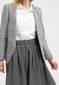 Blazer de patrón de espiga en gris y blanco sobre una camisa blanca de botones. Combinado con una falda gris de talle alto, con pliegues.
