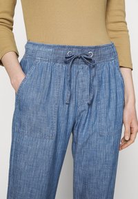 Person som bär högmidjade blå denimbyxor med dragsko i midjan och en långärmad beige topp, med händerna vilande nära fickorna.