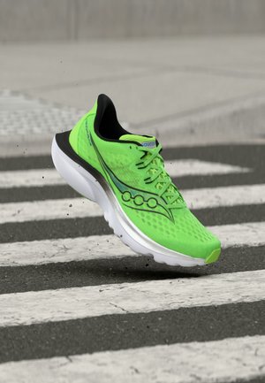 Jasně zelený běžecký boty s černými a bílými detaily, ve vzduchu nad přechodem pro chodce na dlážděné ulici.