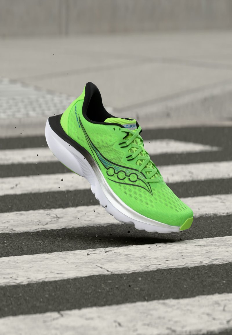 Φωτεινό πράσινο αθλητικό παπούτσι με μαύρες και λευκές λεπτομέρειες, στον αέρα πάνω από διαβάση πεζών σε πλακόστρωτο δρόμο.