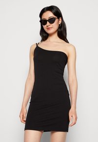 Vestido preto de um ombro só feito de tecido elástico, design justo, até ao joelho, com alças finas e uma textura suave. Usado com óculos de sol estilo gato.