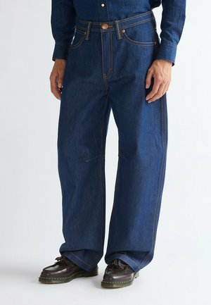 Personne portant un jean bleu foncé à jambes larges avec des coutures contrastées et des chaussures en cuir marron foncé à lacets sur un fond clair.