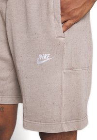 Main dans la poche d'un short Nike beige tacheté avec un logo blanc, montrant une partie d'une jambe tatouée.
