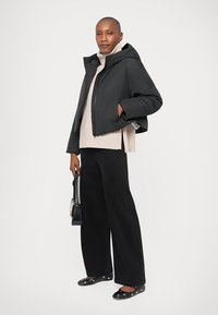 Mujer que lleva una chaqueta de plumas negra con capucha, un suéter de cuello alto beige, pantalones anchos negros, zapatos planos negros con detalles de ojetes y sosteniendo un pequeño bolso negro.