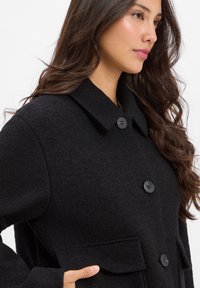 Black felt coat featuring a collared neckline, two front pockets, and three black buttons for closure. Soft texture, loose-fit design.