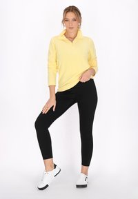 Gul pullover med lynlåskrave, parret med sorte leggings og hvide sneakers med sorte detaljer. Glat tekstur, tætsiddende design.