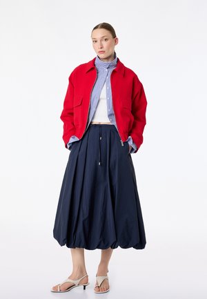 Kobieta stoi z rękami w kieszeniach, ubrana w czerwoną kurtkę, niebieską pasiastą koszulę, biały krótki top, granatową spódnicę balonową oraz białe sandały na niskim obcasie.