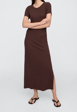 Mujer con vestido maxi marrón de manga corta y abertura lateral, y sandalias planas negras, de pie con los brazos cruzados frente a un fondo liso.