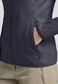 Giacca a zip grigio scuro con tessuto texture, dotata di tasche laterali e orlo con cordoncino. Design aderente con pantaloni beige a contrasto.