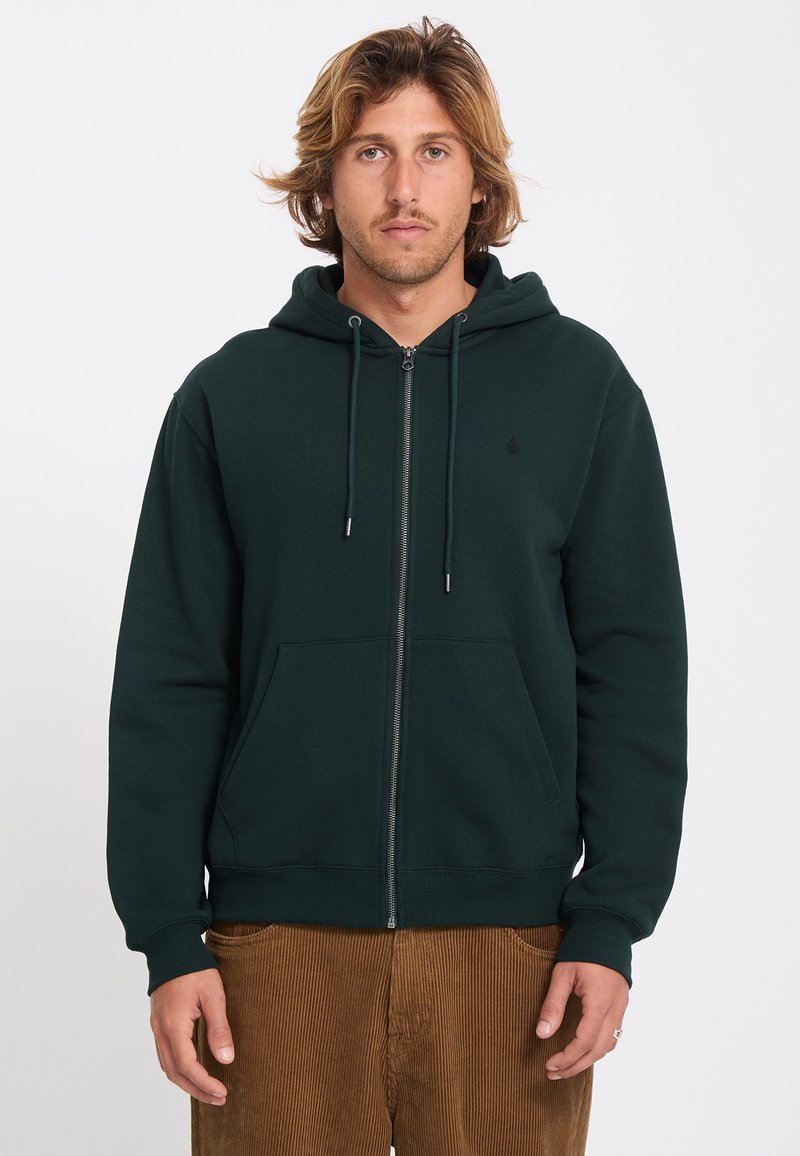 Homme aux cheveux blonds mi-longs porte un sweat à capuche zippé vert foncé et un pantalon en velours côtelé marron, debout devant un fond uni.