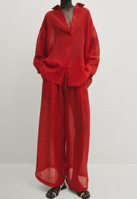Chemise rouge transparente à boutons avec col ouvert, associée à un pantalon assorti à jambes larges. Tissu léger et texturé avec une coupe ample.