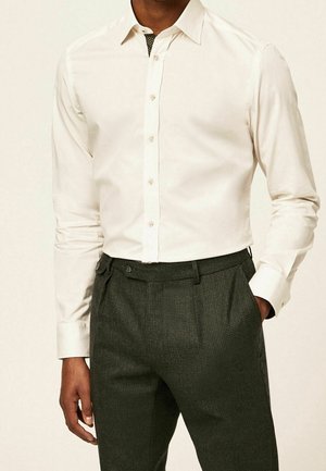 Man wearing a long-sleeve white button-up shirt tucked into dark gray textured dress pants, standing with one hand in pocket.