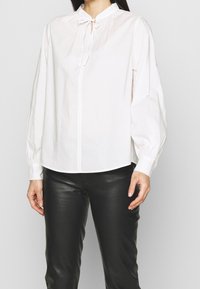 Kobieta w białej bluzce z długim rękawem i wiązaniem przy kołnierzyku, w czarnych skórzanych spodniach, stojąca na tle jednolitego tła.