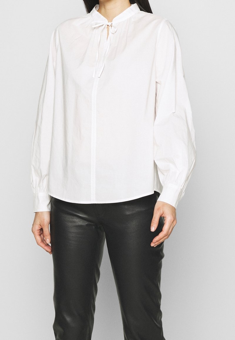 Kobieta w białej bluzce z długim rękawem i wiązaniem przy kołnierzyku, w czarnych skórzanych spodniach, stojąca na tle jednolitego tła.