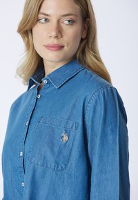 Giovane donna con capelli biondi che indossa una camicia blu con bottoni e un piccolo logo ricamato sulla tasca del petto, che guarda di lato.