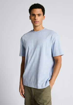 Junger Mann mit kurzen lockigen Haaren, der ein hellblaues kurzärmeliges T-Shirt und olivgrüne Hosen trägt, steht mit einer Hand in der Tasche.