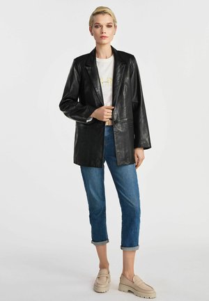 Czarna skórzana marynarka z wyłami, luźny krój, zestawiona z białym t-shirtem i podwiniętymi niebieskimi dżinsami, dopełniona beżowymi butami wsuwanymi.