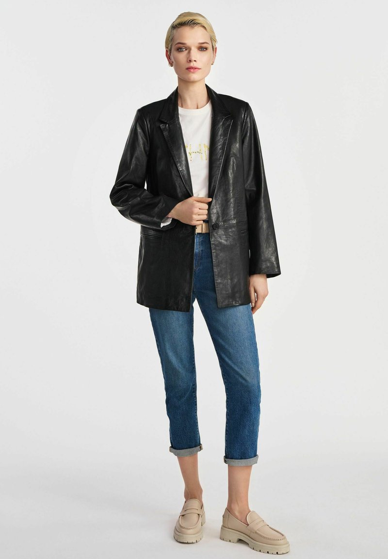 Czarna skórzana marynarka z wyłami, luźny krój, zestawiona z białym t-shirtem i podwiniętymi niebieskimi dżinsami, dopełniona beżowymi butami wsuwanymi.