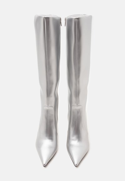 Paire de bottes argentées brillantes montantes jusqu’au genou avec bouts pointus et fermetures éclair au dos, debout sur un fond uni clair.