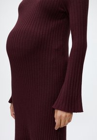 Personne enceinte portant une robe tricotée bordeaux à manches longues côtelées, montrant un gros plan du ventre de grossesse et du bras.
