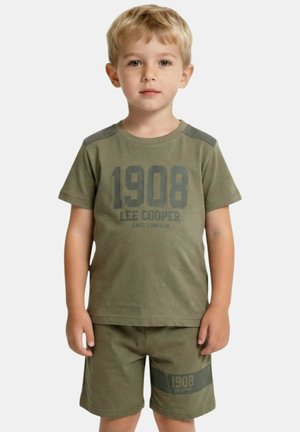 Garçon blond debout, portant un ensemble vert Lee Cooper avec "1908" imprimé sur le t-shirt et le short, sur un fond clair uni.