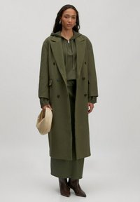 Olivgrüner Übermaßmantel mit Knöpfen, kombiniert mit einem passenden Kapuzenpullover und Rock. Das Outfit beinhaltet braune Absatzstiefel und eine cremefarbene Tasche.
