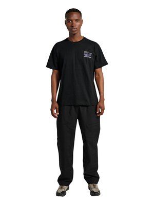 Man staat recht vooruit kijkend, draagt een zwart T-shirt met grafische opdruk, losse zwarte broek en grijze en zwarte sneakers op een witte achtergrond.