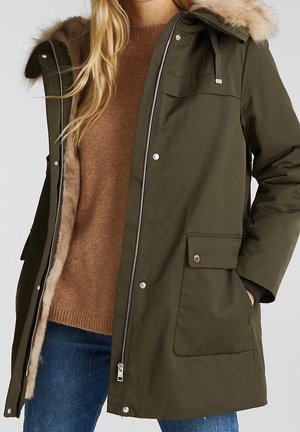 Femme portant un parka vert olive avec une capuche bordée de fourrure, ouvert pour montrer un pull marron et un jean bleu.